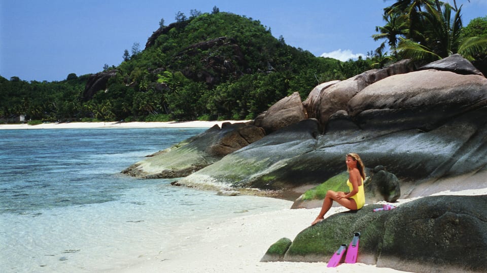 A woman sitting on a large rock by a beach with clear turquoise water and white sand.
