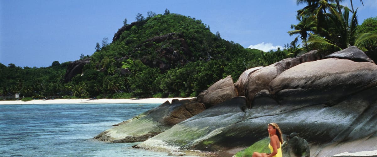 A woman sitting on a large rock by a beach with clear turquoise water and white sand.