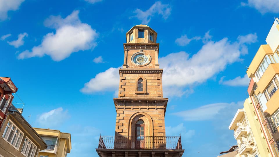 A tall clock tower with a balcony, surrounded by buildings under a clear blue sky.