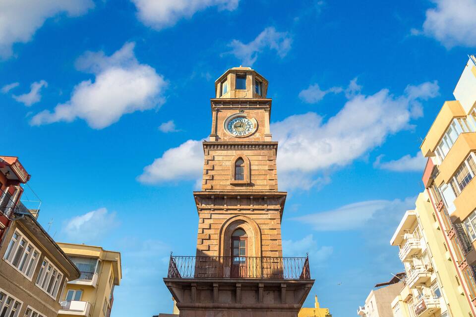 A tall clock tower with a balcony, surrounded by buildings under a clear blue sky.