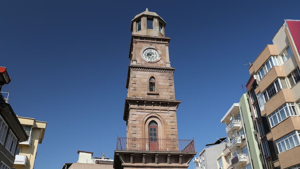 A tall clock tower with a balcony, surrounded by modern buildings.