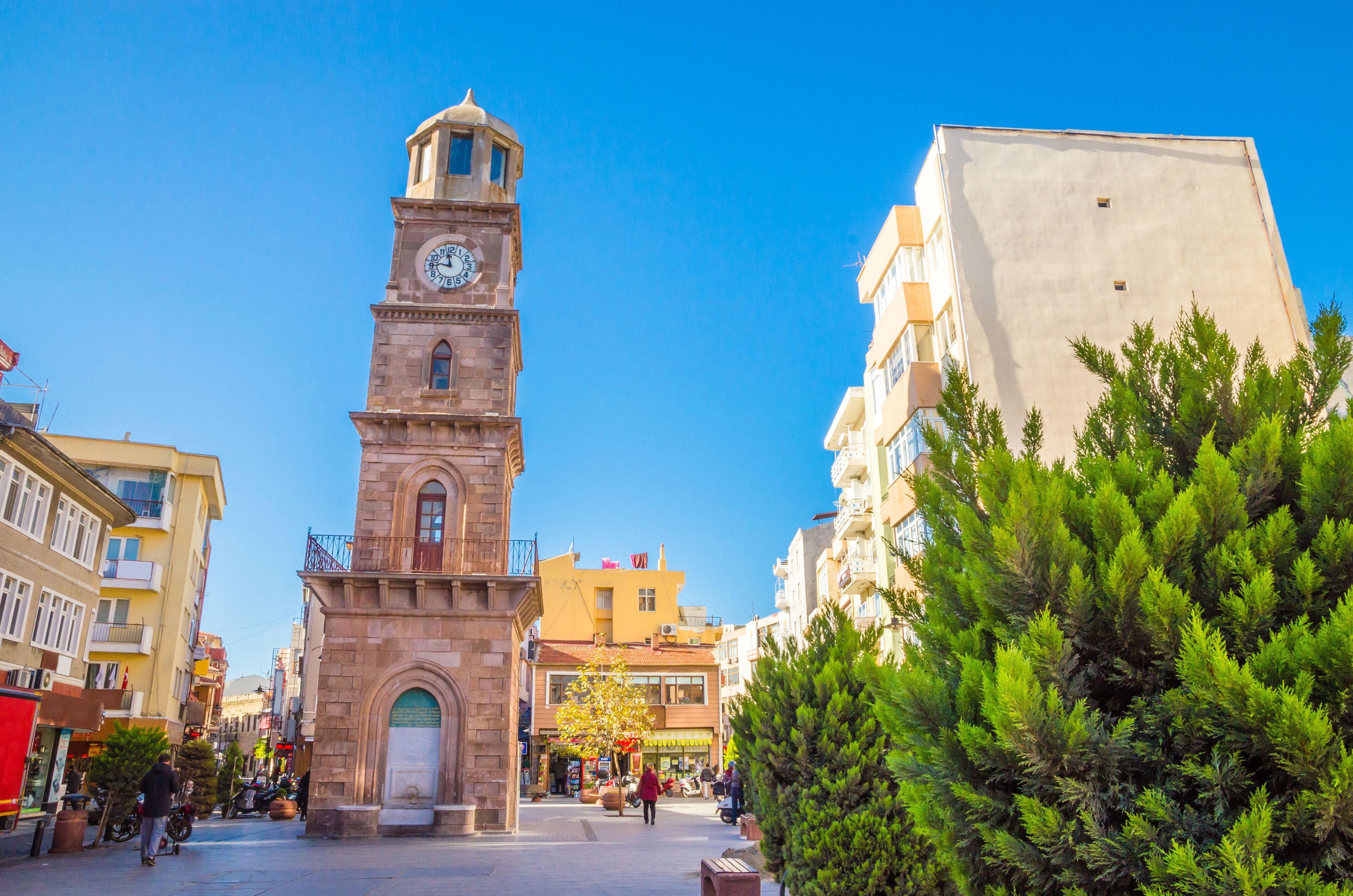 A historic clock tower, a tree-lined square, and modern buildings.