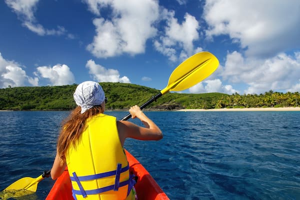 Une personne pratiquant le kayak par une journée ensoleillée, avec une plage et des collines en arrière-plan.