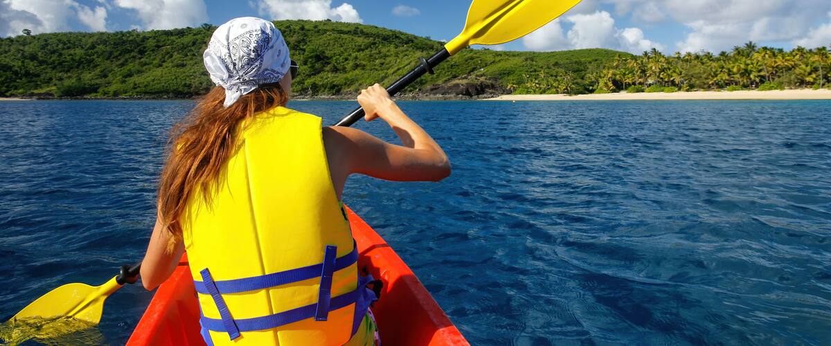 A person kayaking on a clear day with a beach and hills in the background.
