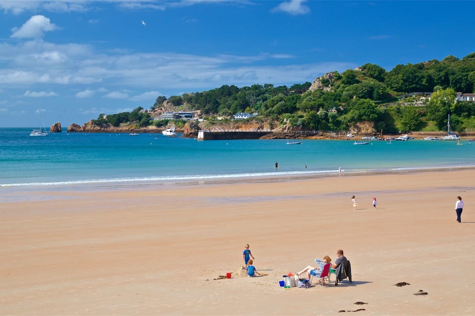 A sandy beach with a clear blue sea, boats, and houses on a hillside in the background.