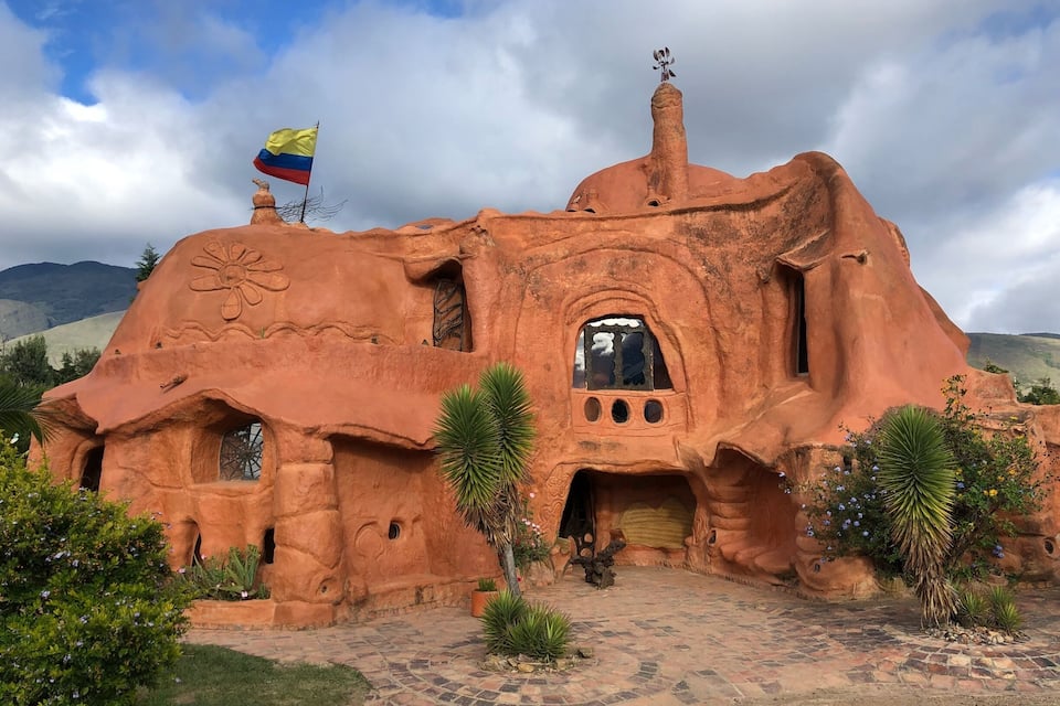 A unique, earth-toned building with a flag, surrounded by greenery and a mountainous landscape.