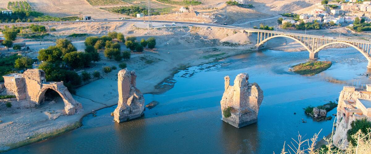 A river with stone arches, a bridge, and a town in the background.