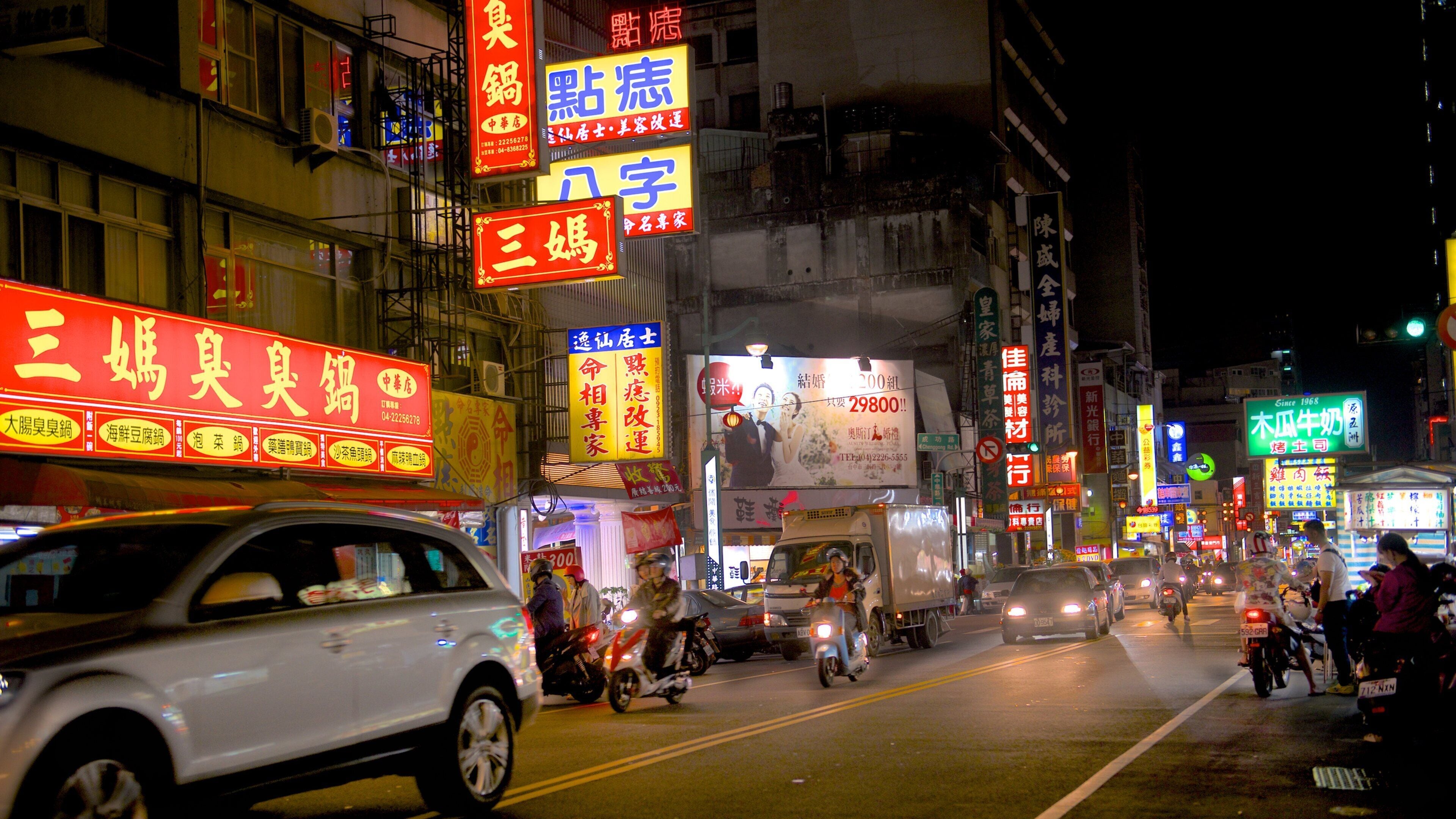 A busy street at night with neon signs in a foreign language, cars, and motorcycles.