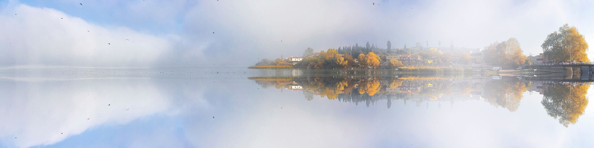 Un lago brumoso con árboles otoñales reflejados en el agua, un puente y una ciudad lejana al fondo.