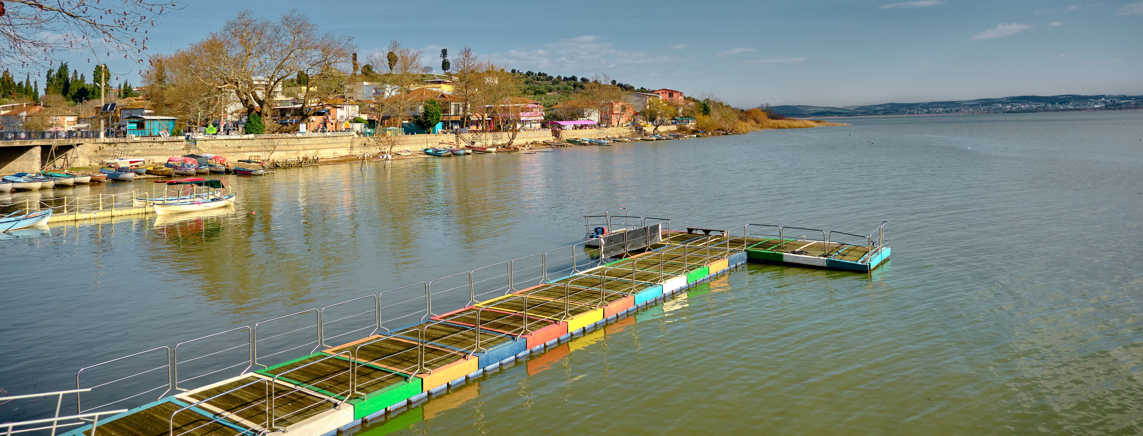A riverside town with boats docked along the shore, a pedestrian bridge, and buildings on a hillside.
