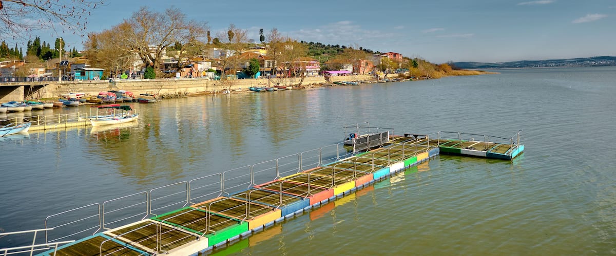A riverside town with boats docked along the shore, a pedestrian bridge, and buildings on a hillside.
