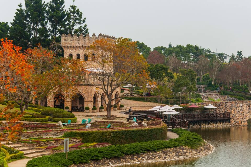 A historic castle with a moat, surrounded by autumn foliage and a riverside café area.