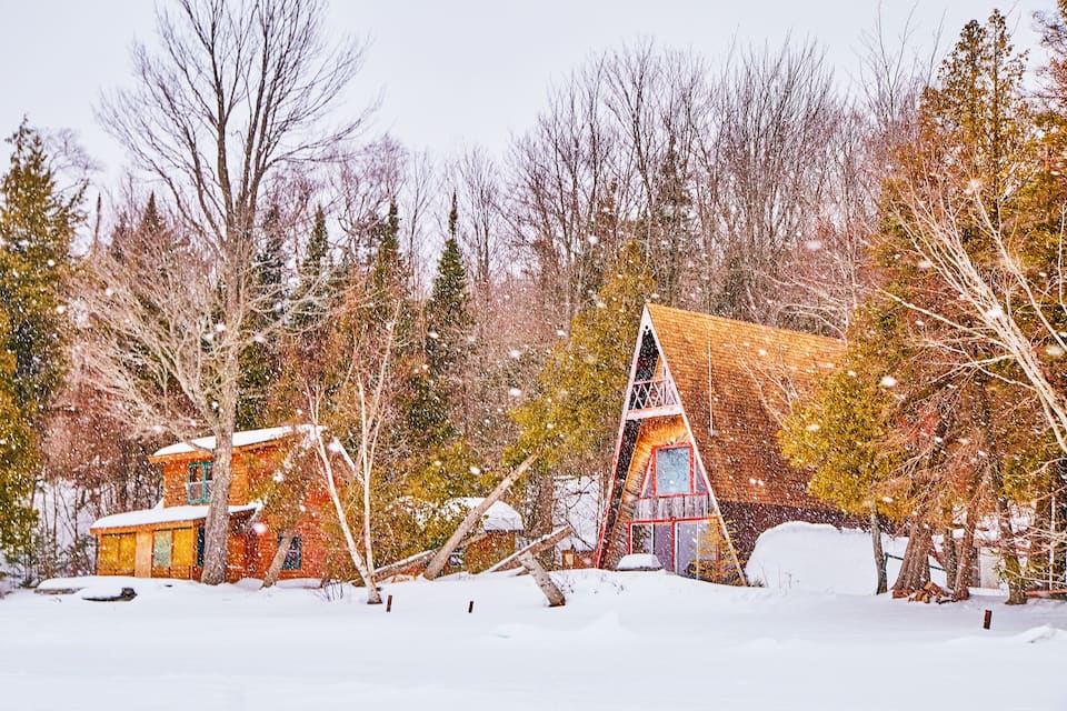 A snow-covered cabin with a red roof and a wooden structure in a forest.