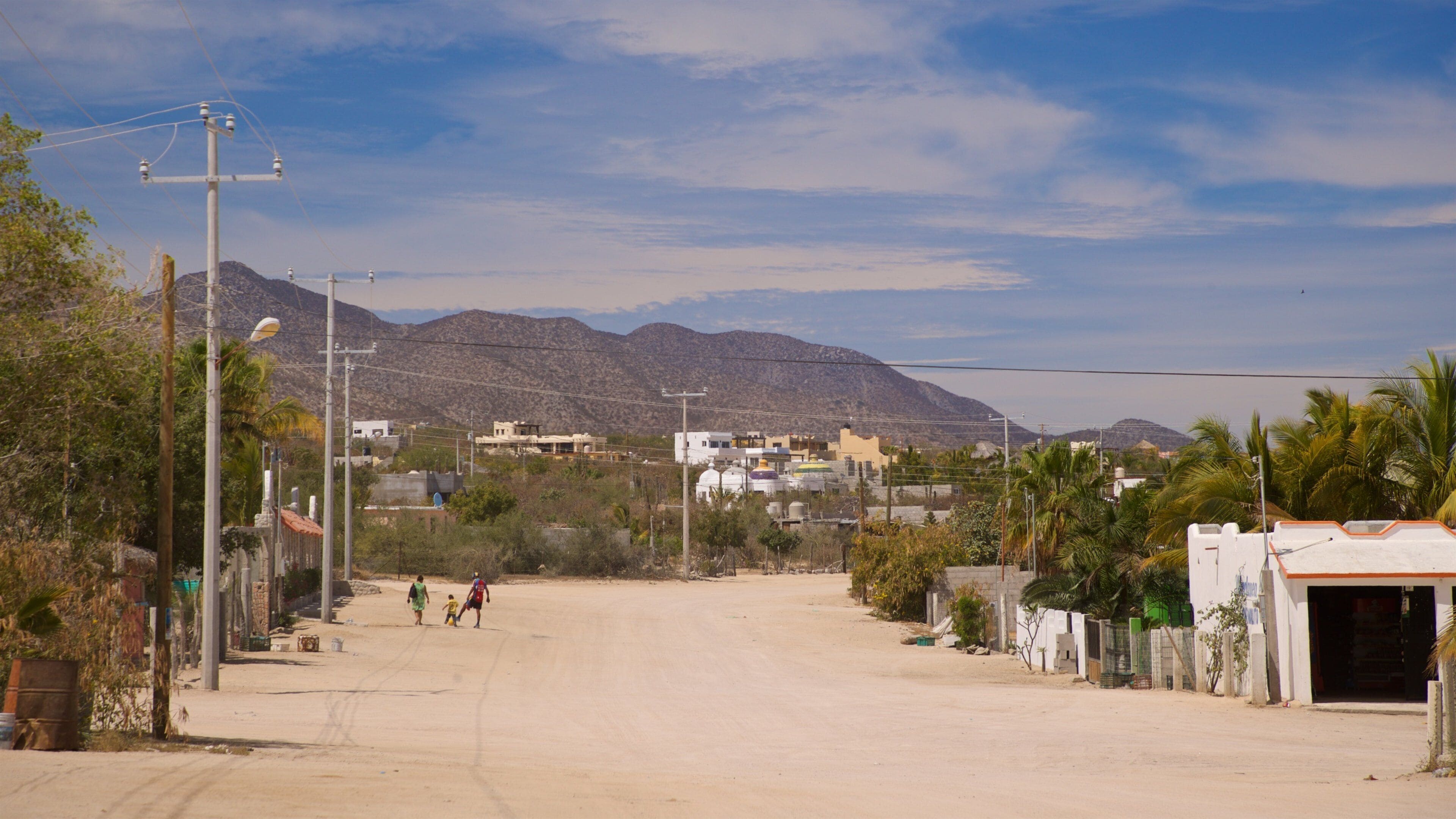 A wide, empty road with mountains in the background, surrounded by sparse vegetation and buildings.