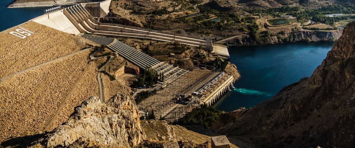 A hydroelectric dam with multiple spillways, a reservoir, and surrounding mountainous terrain.