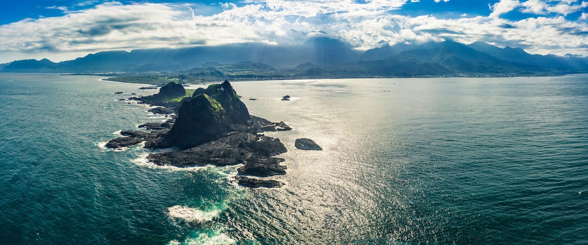 A coastal landscape with a prominent rocky outcrop surrounded by calm sea waters, distant mountains, and a partly cloudy sky.