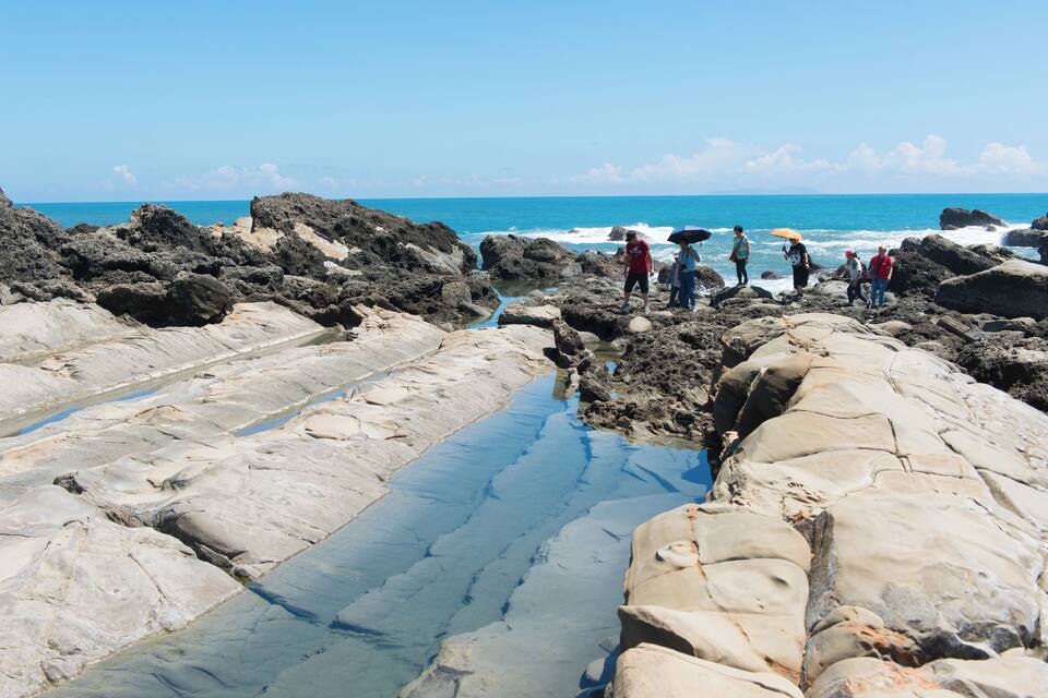 A rocky beach with clear blue water and people standing on the rocks.