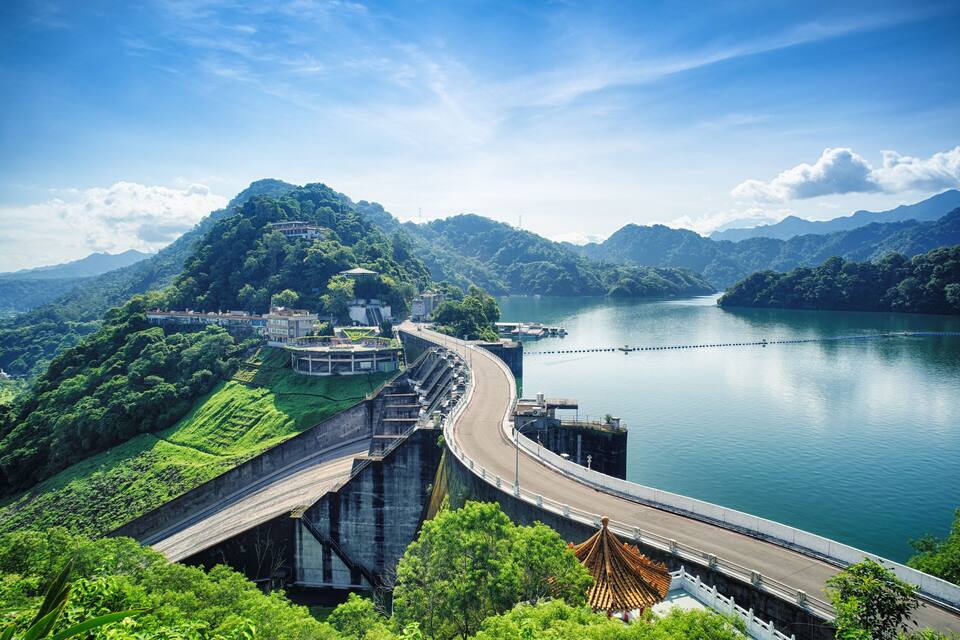 A dam with a road curving over it, surrounded by lush green hills and a calm lake.