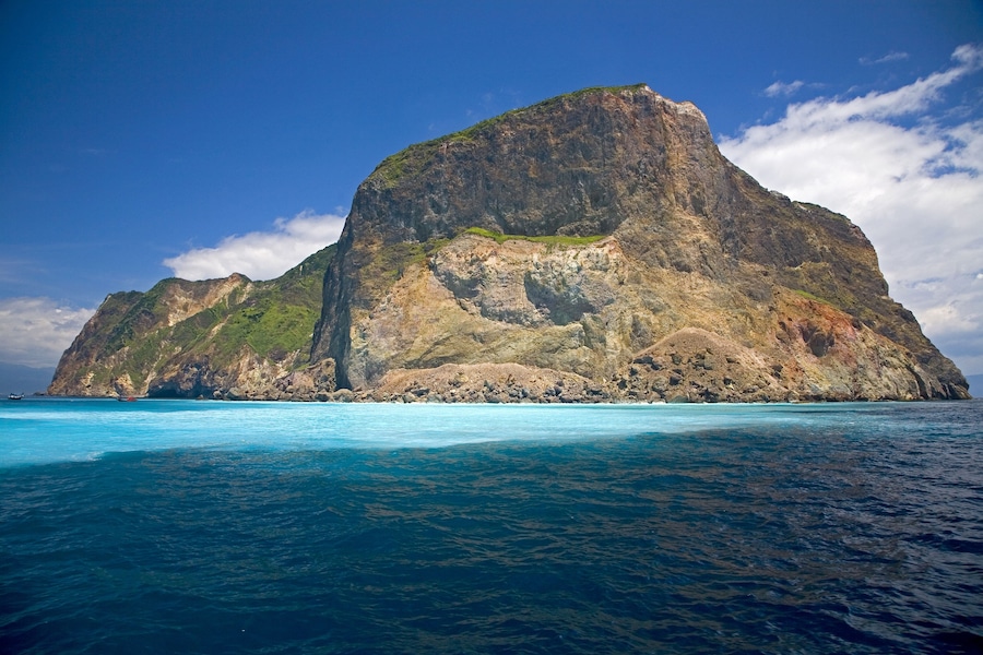 Una isla grande y rocosa con un acantilado empinado y un pequeño parche de vegetación verde en la cima, rodeada de aguas azules cristalinas.