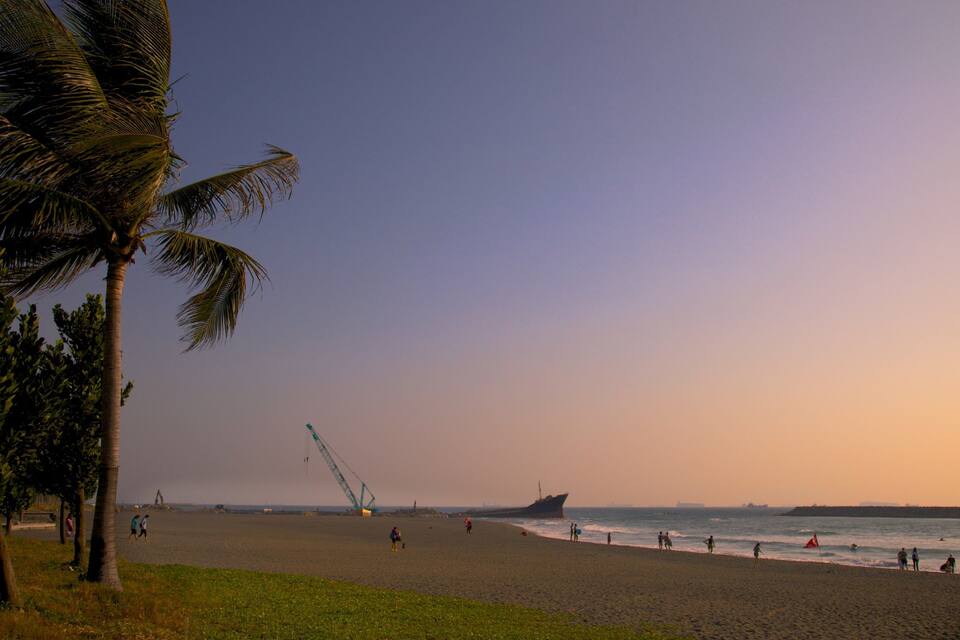 A beach with palm trees, a large ship, and a crane in the distance.