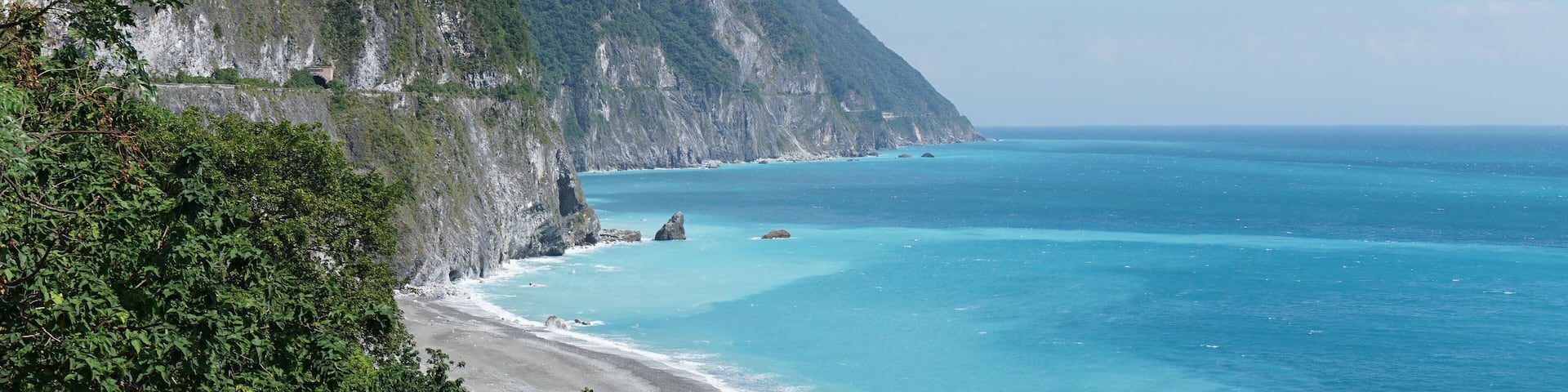 A coastal landscape with a steep cliff, turquoise sea, and dense greenery.