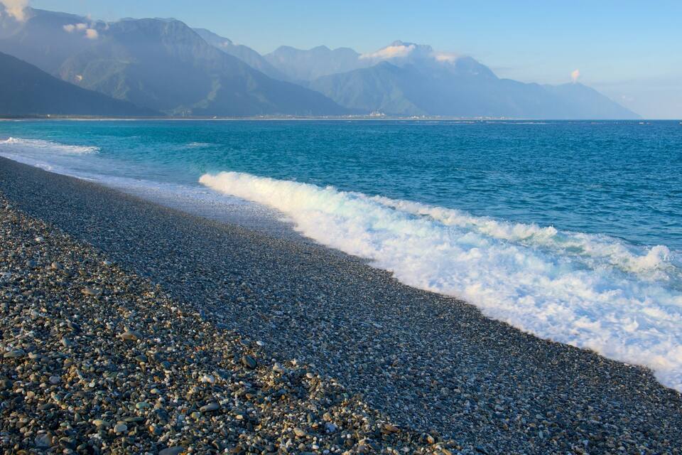 Una playa de guijarros con olas, montañas y cielo despejado.
