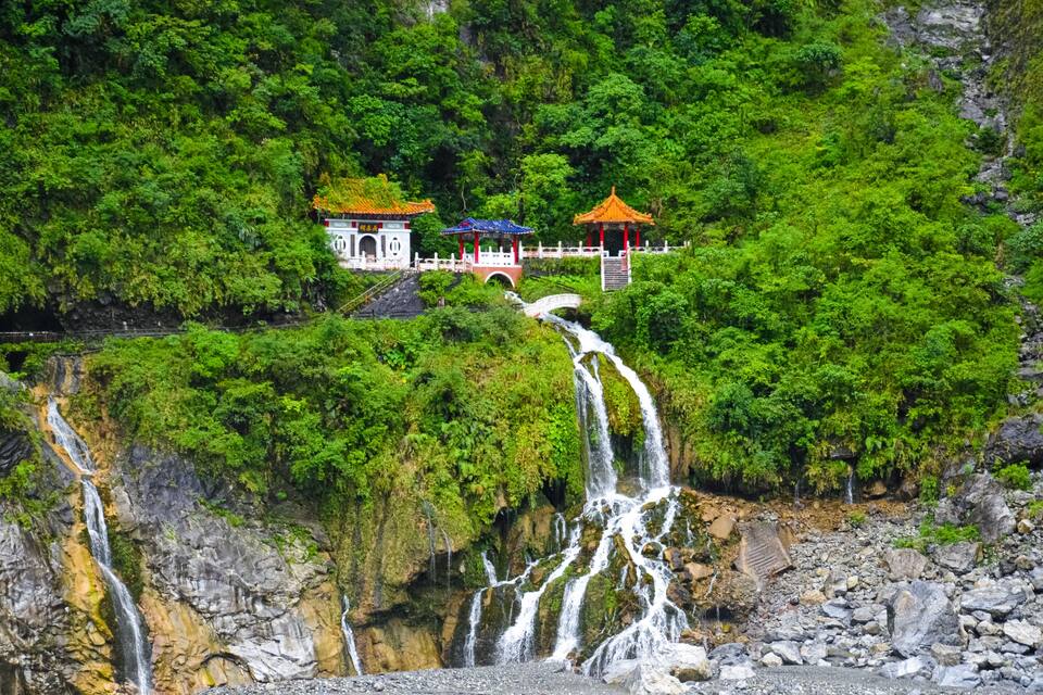 Una cascata si getta da una parete rocciosa, circondata da una vegetazione lussureggiante e da architettura tradizionale cinese.