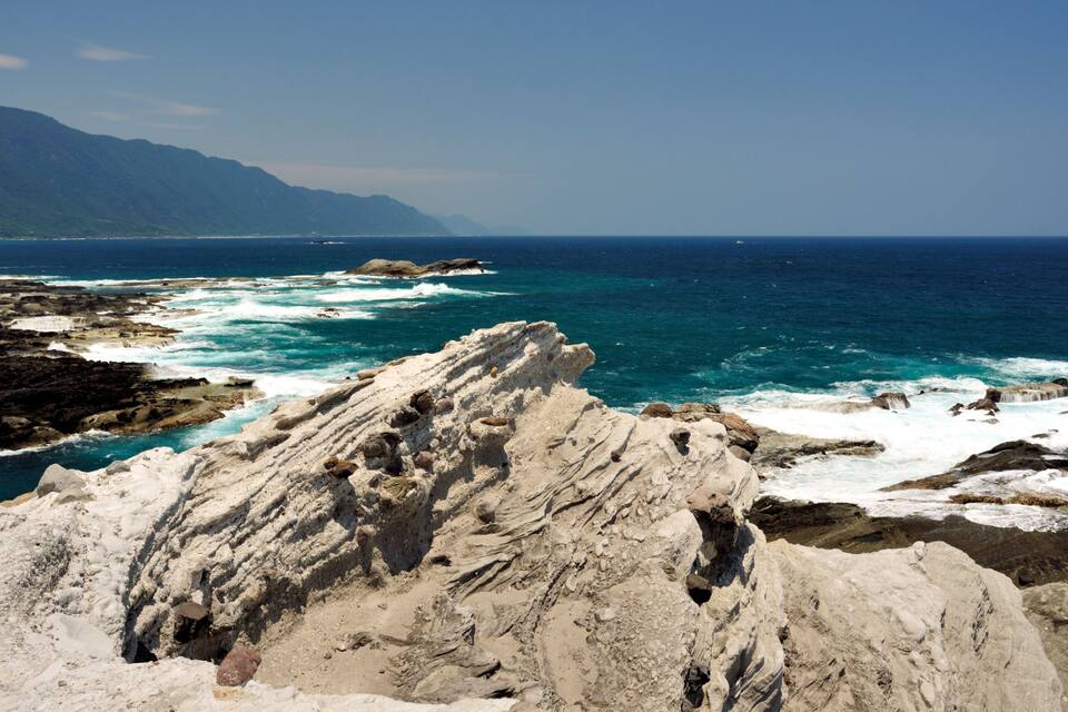 A rocky coastline with waves crashing against the shore and mountains in the background.
