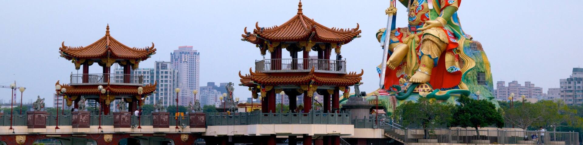 Una estatua imponente de una deidad sobre un puente que cruza el agua, con arquitectura china tradicional al fondo.