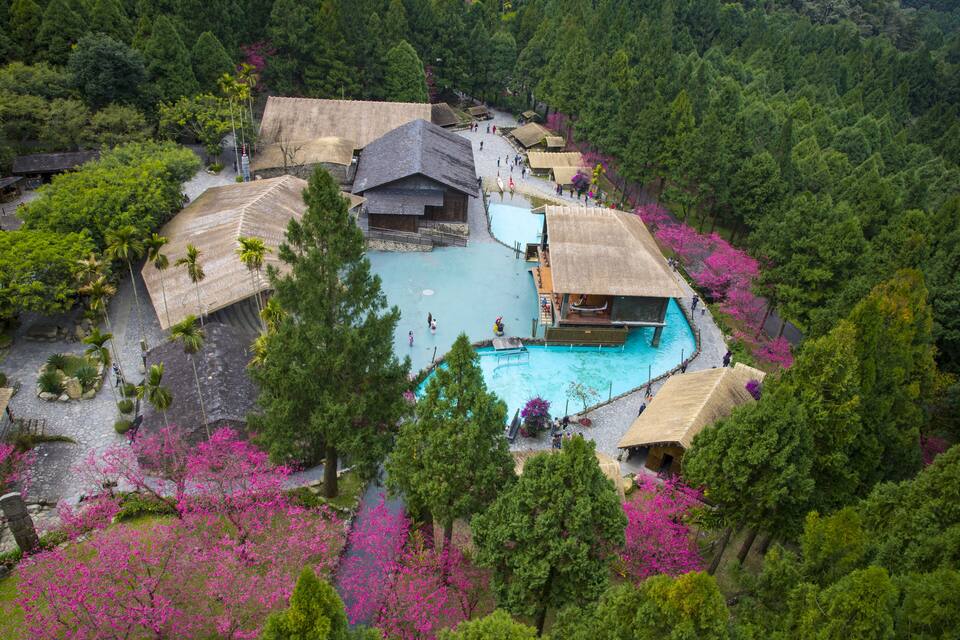 A traditional village with a central swimming pool surrounded by thatched-roof buildings and lush greenery.
