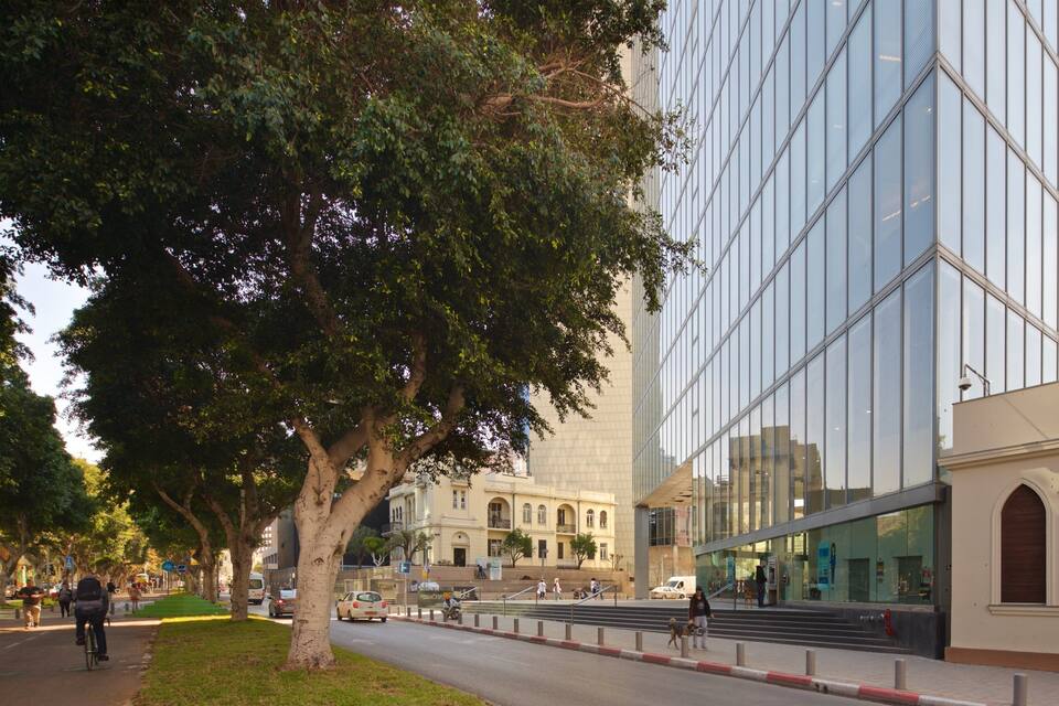 A modern building with reflective glass windows, a tree-lined sidewalk, and a historic building in the background.