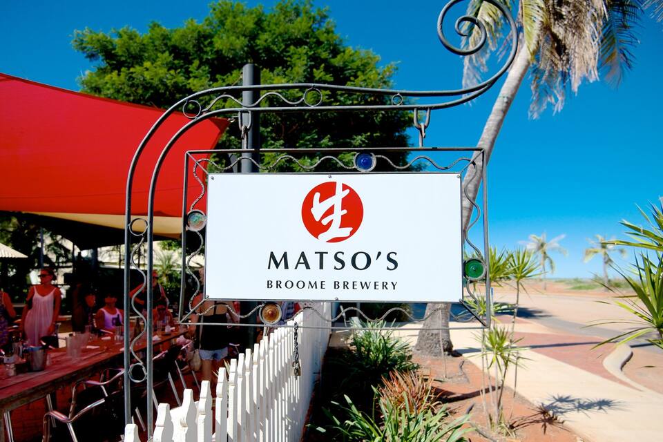 A sign for Matso's Broome Brewery with a red and white logo, surrounded by palm trees and a clear blue sky.
