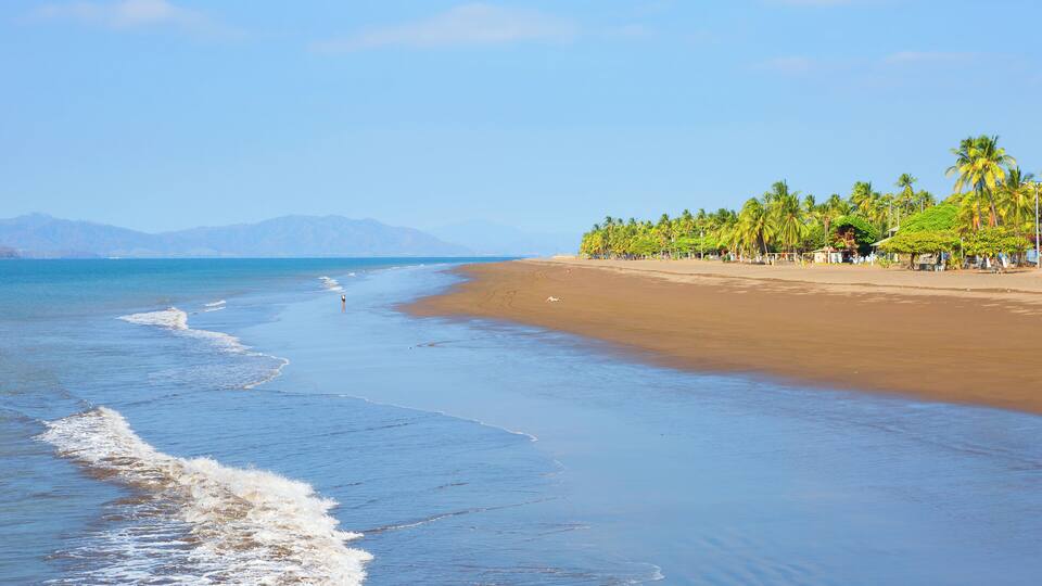 A beach with waves, palm trees, and mountains in the background.