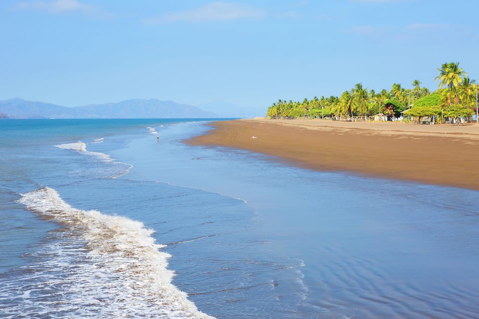 A beach with waves, palm trees, and mountains in the background.