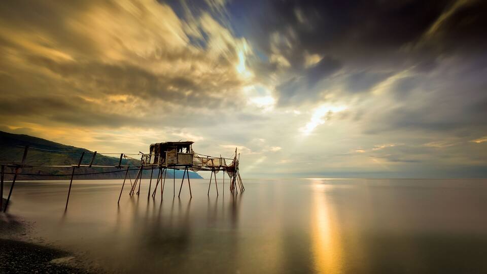 A wooden structure on stilts over calm water, surrounded by a mountainous landscape under a dramatic sky.