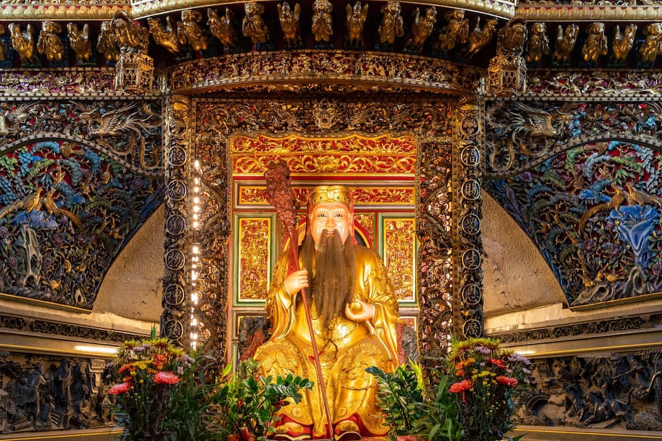 Eine goldene Statue einer Figur mit Stab, umgeben von aufwendigen Schnitzereien und farbenprächtigen Blumenarrangements.