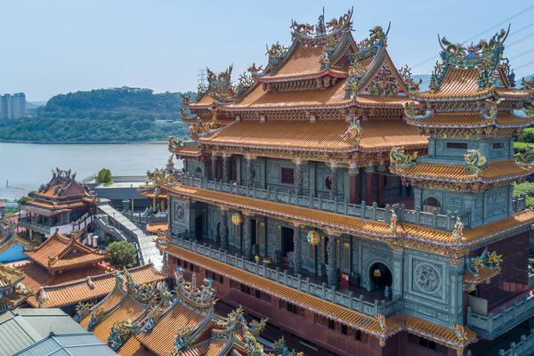 Un temple traditionnel à plusieurs niveaux, doté de toits richement décorés et de sculptures complexes, situé au bord d’un plan d’eau.