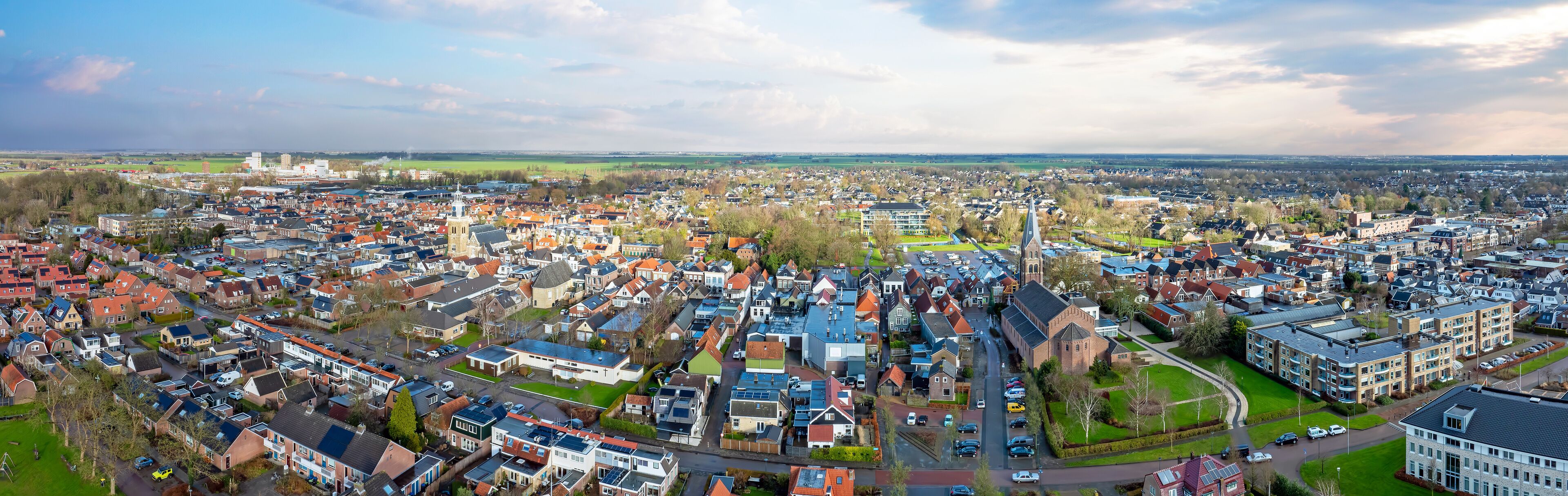 An aerial view of a densely populated urban area with a mix of residential houses and commercial buildings, surrounded by green fields.