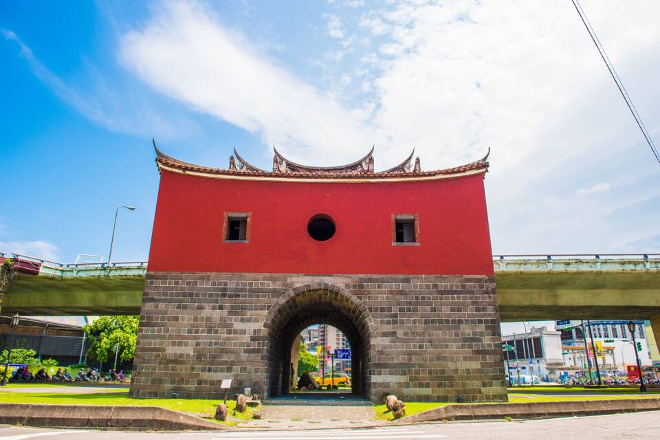 A red and grey archway with a traditional roof, under a bridge.