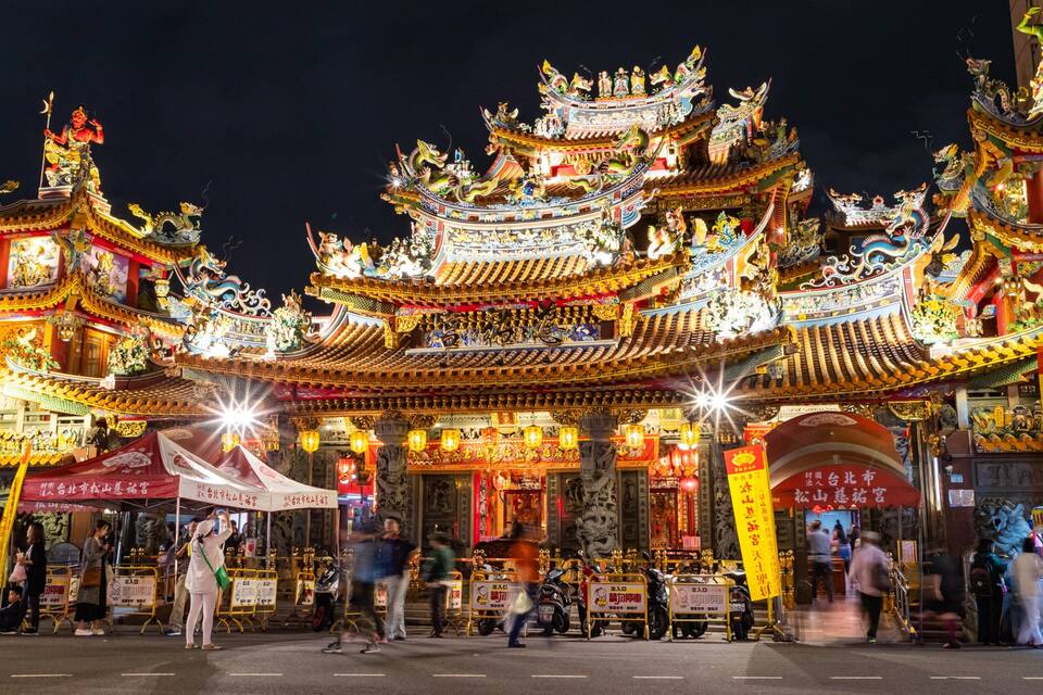 A brightly lit traditional Chinese temple with ornate architecture and a crowd of people in front.