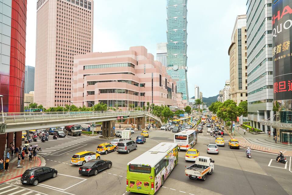 A busy urban street with tall buildings, a pedestrian bridge, and various vehicles including buses, taxis, and cars.