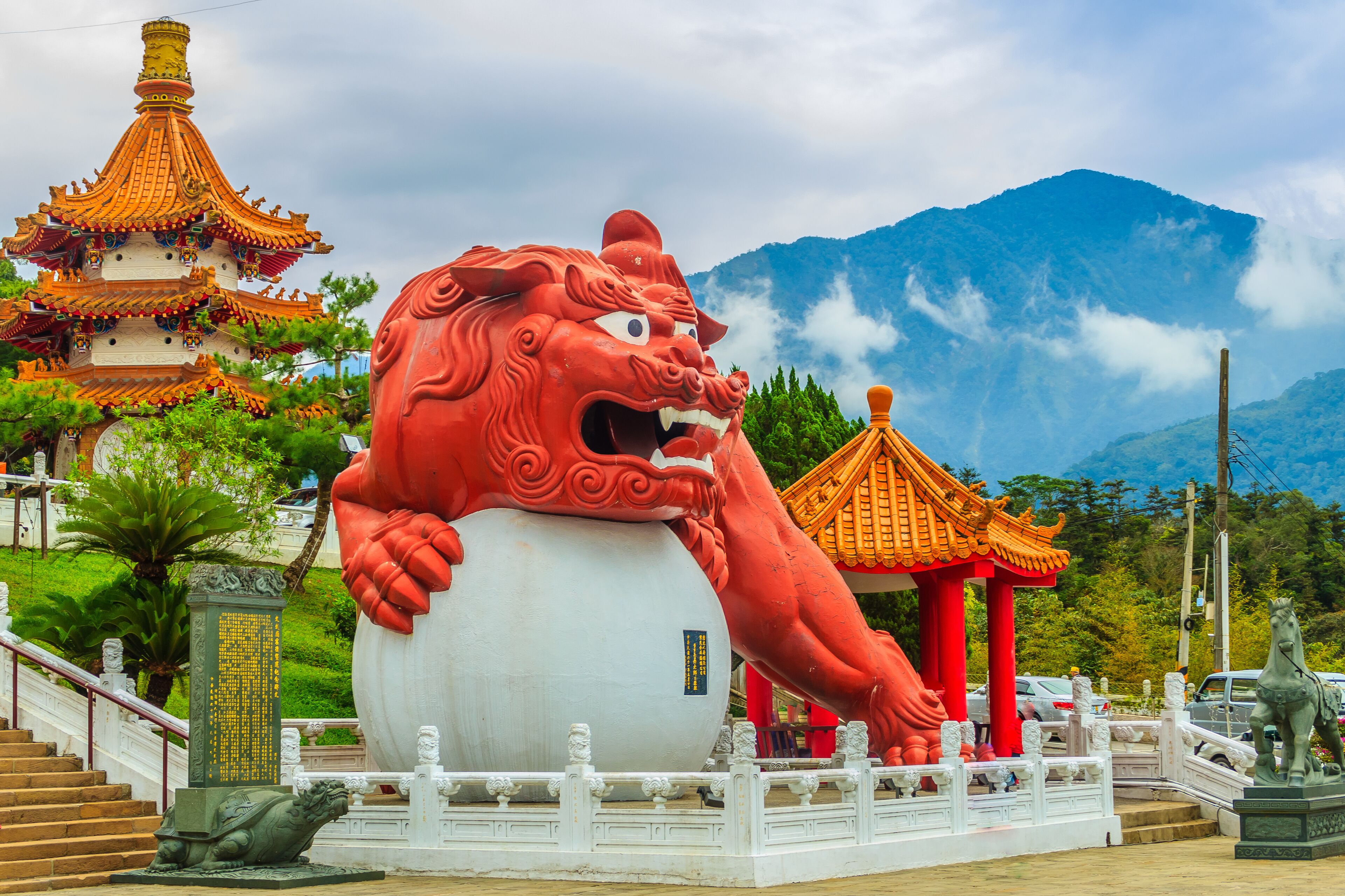 A large red lion statue, a traditional Chinese temple, and a mountain in the background.