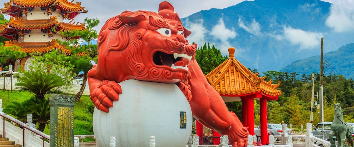 A large red lion statue, a traditional Chinese temple, and a mountain in the background.