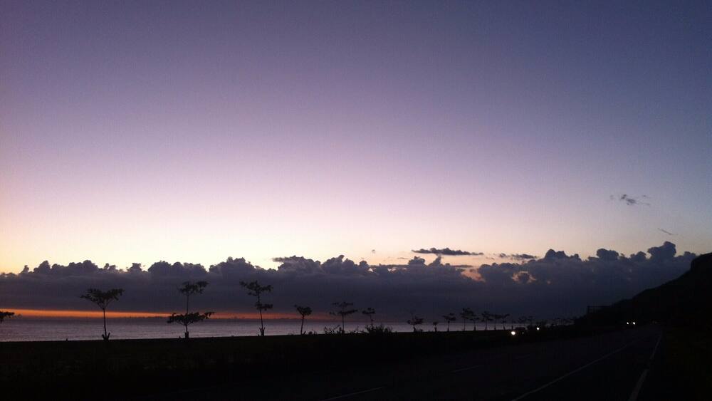A sunset over a coastal road with silhouettes of trees and a distant body of water.