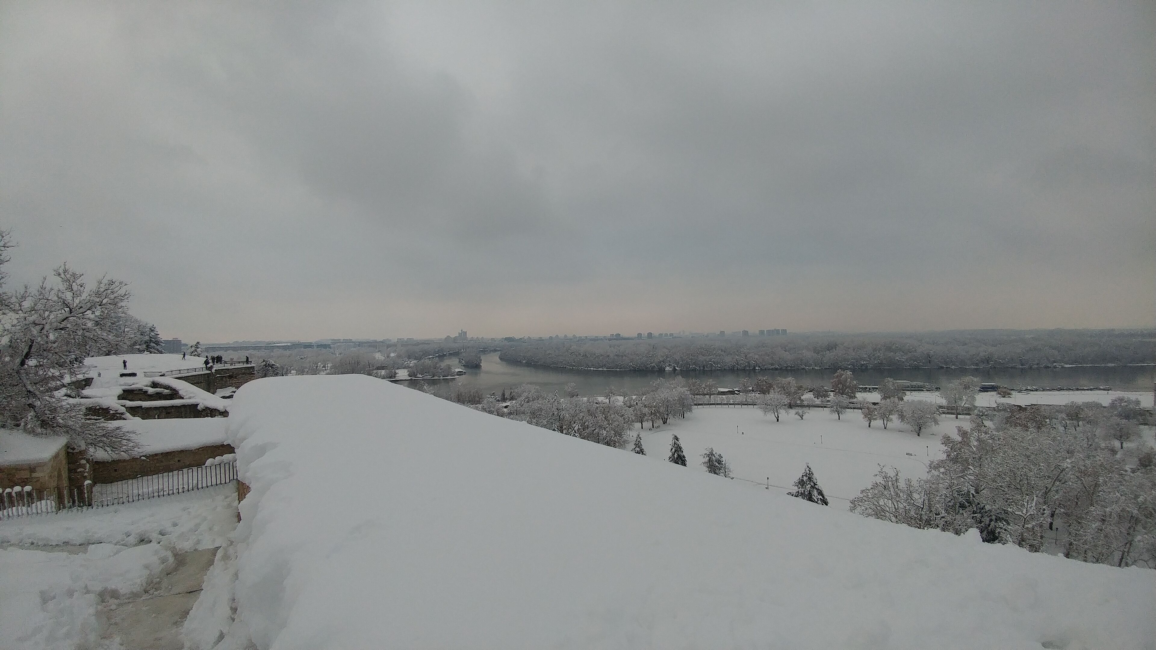 A snow-covered landscape with a river, trees, and a distant city skyline.