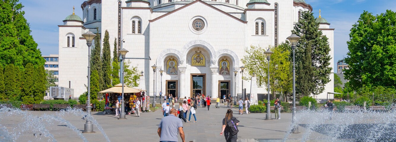 Uma igreja grande e branca com cúpula verde e cruzes douradas, cercada por uma fonte e árvores.
