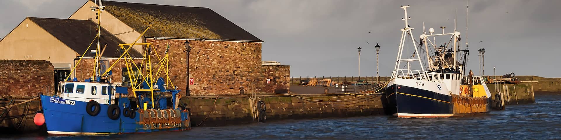 Two boats docked at a harbor with a brick building in the background.