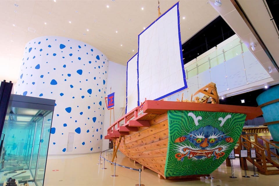 Un modellino di nave in legno con decorazioni a drago, un'ampia parete bianca con macchie azzurre e un edificio moderno con finestre in vetro.