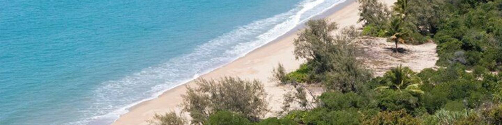 A coastal landscape with a sandy beach, clear blue water, and dense green vegetation.