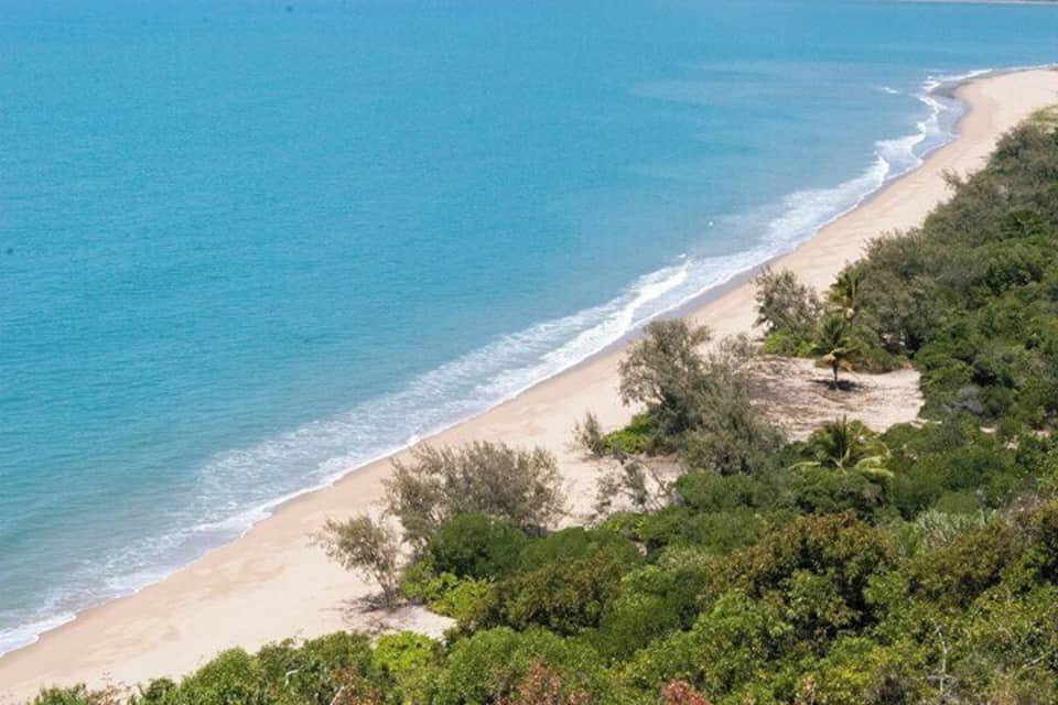 A coastal landscape with a sandy beach, clear blue water, and dense green vegetation.